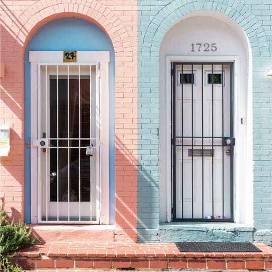Two front doors next to each other. Door on the left surrounded by pink painted bricks while the door on the right is surrounded by light blue painted bricks.