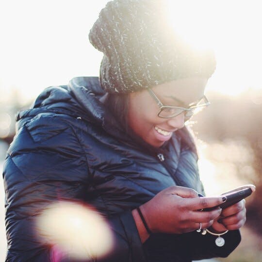 Image of a woman in a winter coat and hat looking down at a mobile phone and smiling. 