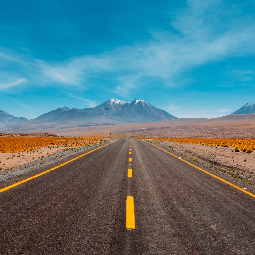 A long, winding highway in the desert without any cars. A snow covered mountain is in the distance. Blue skies with minimal clouds.