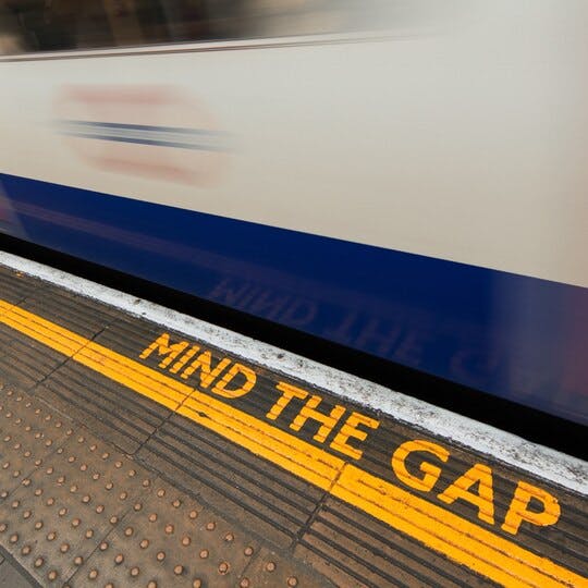 Image of a train going by a platform that says "Mind the Gap" on it.