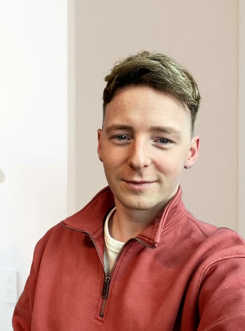 A smiling Emmet O’Flaherty, wearing a reddish-brown zip-up jacket and a white shirt, seated against a beige background.