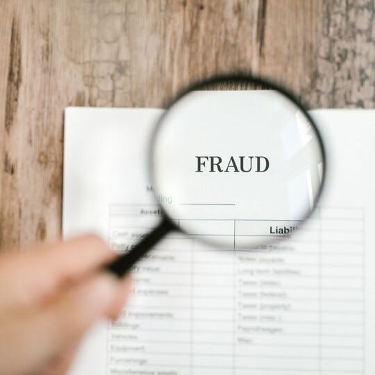Magnifying glass over the word "FRAUD" on a piece of paper lying on a wooden surface.