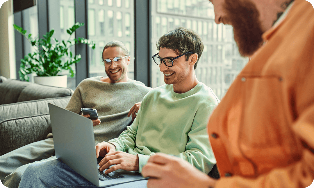 Group of three colleagues sitting on a couch smiling while working together on a laptop and mobile phone.