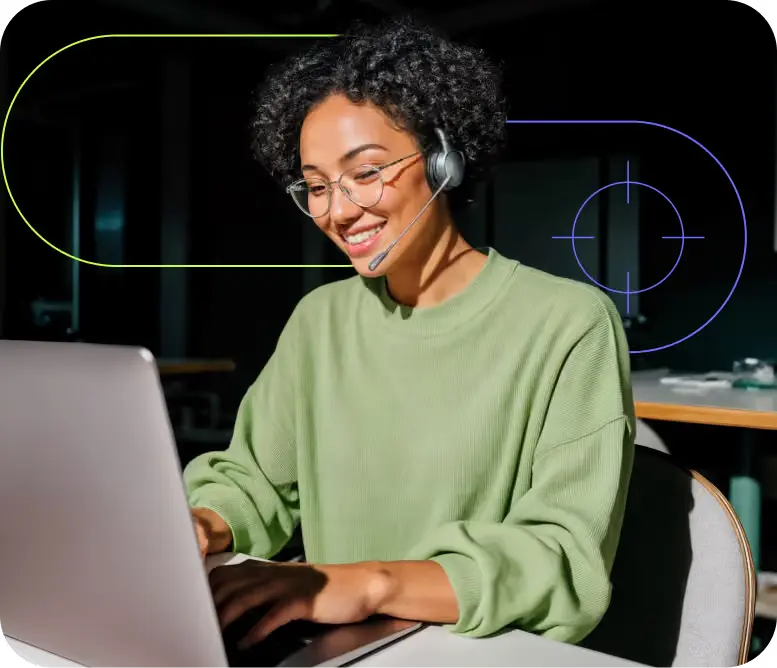 Woman in a headset smiling as she works at a laptop in a dim office